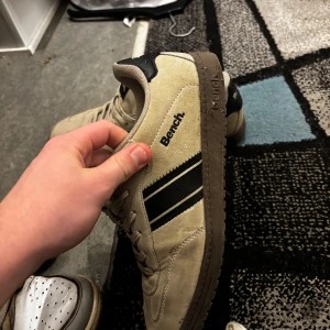 Beige sneakers från Bench - Snygga beige sneakers från Bench med svarta detaljer och två svarta ränder på sidan. Skorna har snörning och en platt brun sula. Materialet är mocka och syntet, vilket ger en cool och stilren look. Perfekta för dig som gillar klassisk streetstil.