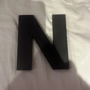 Dekorativt bokstav N i trä - Stilren svart bokstav N i trä, perfekt för dekoration i hemmet eller på kontoret. Bokstaven är i gott skick med mindre spår av användning på baksidan. Kan ställas eller hängas upp. Passar utmärkt som personlig inredningsdetalj.