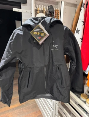 Svart Arc'teryx Gore-Tex vindjacka M - Svart vindjacka från Arc'teryx i storlek M, tillverkad i slitstark Gore-Tex. Jackan har huva, flera dragkedjeförsedda fickor och minimalistisk design med Arc'teryx-logga på bröstet. Perfekt för dig som gillar outdoor och vill ha en clean look.