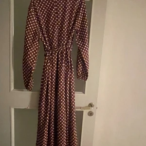 Brun lång prickig klänning med bälte - Lång brun klänning med vita prickar och matchande bälte i midjan. Klänningen har långa ärmar med resår vid handlederna och en avslappnad passform. Perfekt för dig som gillar retrovibbar och mönster som sticker ut.