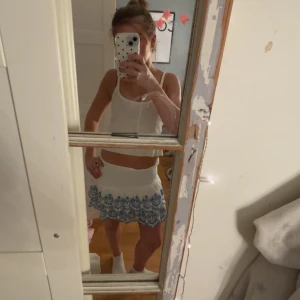 Vit kortkjol med blått mönster - Supersöt vit kortkjol med blått broderat mönster längs nederkanten. Kjolen har en lätt volang och sitter snyggt i midjan.