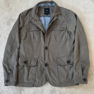 Grå vindjacka från Riley, storlek 50 (M) - Snygg grå vindjacka från Riley med klassisk krage och flera fickor med lock framtill. Jackan har både dragkedja och knappar, vilket ger en cool look. Materialet är lätt och smidigt, perfekt för dig som gillar stilren design och funktion. Modellen är 184 cm och väger cirka 67kg 