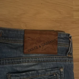 Dolce & Gabbana blå jeans med slitningar - Snygga blå jeans från Dolce & Gabbana med coola slitningar och tvättade detaljer. Klassisk femficksmodell med silverfärgade knappar och dragkedja. Märkespatch i brunt skinn baktill. Passar dig som gillar en avslappnad och trendig look.