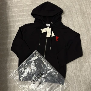 Svart hoodie från Ami med rött hjärta - Svart hoodie från Ami Alexandre Mattiussi med dragkedja, huva och snörning. På bröstet sitter ett broderat rött hjärta med A. Tröjan är i mjuk bomull och har långa ärmar. Perfekt för dig som gillar stilrena detaljer och streetwear.