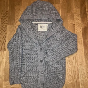 Grå stickad kofta/cardigan - Mysig grå stickad kofta-cardigan från Hudson & Rose med huva och stora knappar framtill. Koftan har lång ärm, ribbade muddar och ett grovt stickat mönster som ger en cool och avslappnad vibe. Perfekt för lager-på-lager under kyliga dagar. Pris går att diskutera vid snabb affär