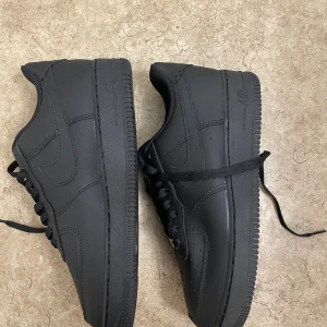 Svarta Nike Air Force 1 sneakers - Säljer ett par helsvarta Nike Air Force 1 sneakers med klassisk låg siluett. Skorna har snörning, perforerad tå och platt sula med tydlig Air-dämpning. Materialet är skinn och detaljerna är ton-i-ton, vilket ger en clean och stilren vibe.