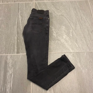 Svarta jeans från Nudie Jeans, 28/32 - Säljer ett par svarta jeans från Nudie Jeans i storlek 28/32. Jeansen har klassisk femficksdesign, svarta sömmar och Nudie-logga på bakfickan. Tillverkade i mjukt denimtyg med en rak passform som sitter snyggt på benen.