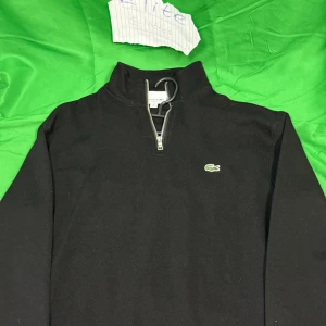 Svart half zip tröja från Lacoste - Svart långärmad tröja från Lacoste med klassisk krokodil-logga på bröstet. Tröjan har en snygg half zip-dragkedja och hög krage. Perfekt för dig som gillar stilren och enkel design med sportig touch.Liknande modell som originalet!