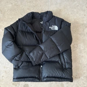 Svart pufferjacka från The North Face - Säljer en svart pufferjacka från The North Face med broderad logga på bröstet och ryggen. Jackan har hög krage, dragkedja framtill och två sidofickor. Stl 154 Perfekt för kalla dagar och riktigt snygg till streetwear-stilen.
