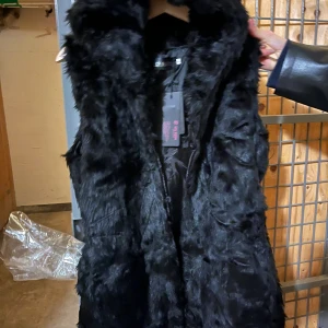 Svart pälsväst från Jen Long - Snygg svart pälsväst från Jen Long med fluffig känsla och hög krage. Västen har en stilren look och är fodrad på insidan för extra komfort. Perfekt att slänga över en hoodie eller långärmad tröja för en cool vibe.