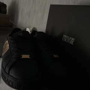 Svarta sneakers Versace Jeans Couture - Svarta sneakers från Versace Jeans Couture med gulddetaljer och logga på sidan samt hälen. Skorna har snörning och är tillverkade i ett strukturerat syntetmaterial med platt sula. Perfekta för dig som vill sticka ut med en lyxig touch.