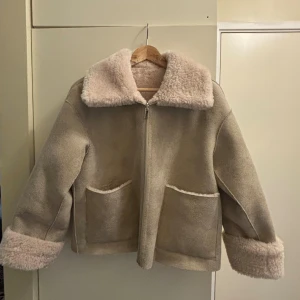 Beige teddyjacka med dragkedja - Säljer en beige teddyjacka med fluffig krage och muddar. Jackan har två stora fickor framtill och stängs med dragkedja. Materialet är mjukt och har en lyxig teddylook, perfekt för kalla dagar. Jackan har en loose passform och är riktigt cozy.