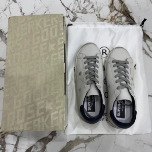 Golden Goose Super-Star sneakers vit/grå - Golden Goose Super-Star sneakers i vitt skinn med grå stjärndetalj på sidan, grå snörning och mörkblå häl. Klassisk rund tå och platt sula med vintage-inspirerade detaljer. Perfekta för dig som gillar en clean men edgy stil.