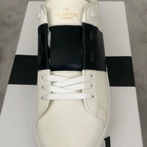 Valentino Garavani Open sneakers vit/svart - Säljer ett par Valentino Garavani Open sneakers i vitt skinn med bred svart läderdetalj över mitten. Klassisk rund tå, vita snören och chunky vit sula med mönstrad undersida. Snygg och stilren design, perfekt för dig som gillar lyxiga streetwear-vibbar.