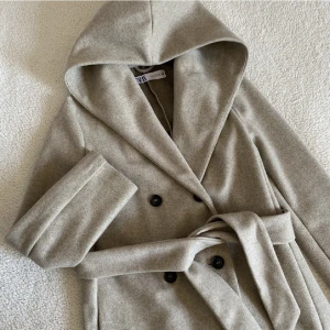 ZARA BEIGE KAPPA MED STOR LUVA - Supergullig kappa med en STOR luva från ZARA. Svarta knappar och bälte för en smalare passform. Endast använd EN GÅNG. Kan förekomma någon liten noppra men annars är den i nyskick.