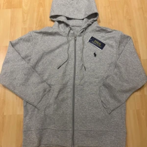 Grå hoodie från Polo Ralph Lauren - Snygg grå hoodie från Polo Ralph Lauren med dragkedja och huva. Klassisk logga broderad på bröstet och praktiska fickor framtill. Tillverkad i mjuk bomullsmix, perfekt för chill dagar. Justerbar snörning i huvan och stilren design.