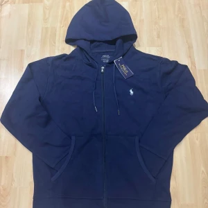 Marinblå hoodie från Polo Ralph Lauren - Snygg marinblå hoodie från Polo Ralph Lauren med dragkedja och huva. Tröjan har snörning vid huvan, två fickor framtill och den klassiska loggan broderad på bröstet. Perfekt för en chill och stilren look.