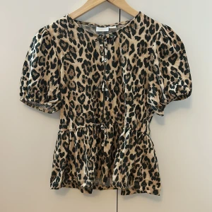 Leopardmönstrad blus från Gina Tricot - Säljer en snygg blus från Gina Tricot med leopardmönster i beige och svart. Blusen har puffiga korta ärmar, rund hals och två knytband framtill. Den har en peplumformad nederdel som ger en cool siluett. Perfekt för dig som gillar att sticka ut!