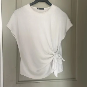 Vit topp med knyt från Zara - Säljer en stilren vit topp från Zara med korta ärmar och snygg knytdetalj i sidan. Toppen har en rund halsringning och är tillverkad i ett mjukt material som känns skönt mot huden. Perfekt för en enkel men trendig look.