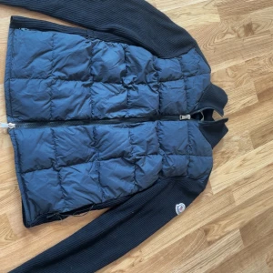 Moncler cardigan  - Snygg svart pufferjacka från Moncler med quiltad front och ribbstickade ärmar och rygg. Jackan har dragkedja framtill och klassisk Moncler-logga på ärmen. Perfekt för dig som vill ha en stilren och exklusiv look under höst och vinter. Båda fickorna gick sönder men har fixat en men en är Forf sönder. Lite nopprig men inget om märks, köpt för 4 år sen