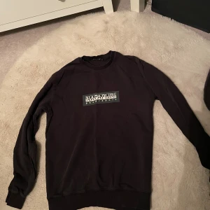 Svart sweatshirt från Napapijri - Svart sweatshirt från Napapijri med stort vitt tryck på bröstet. Tröjan har rund hals, ribbade muddar och lång ärm. Tröjan är lite smått urtvättad men inget som märks stort. Man ser tecken på användning men är fortfarande i gott skick. Säljes för att den blivit för liten. Hör av dig vid fler frågor och funderingar☺️