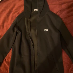 Svart hoodie från Lacoste med dragkedja. Om du är 170 cm+ kommer den passa perfekt  - Svart hoodie från Lacoste med klassisk krokodillogga på bröstet. Tröjan har huva, dragkedja framtill och två stora fickor. Perfekt för dig som gillar en stilren och sportig look.