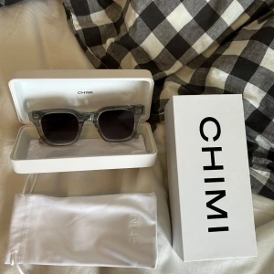 Grå transparenta solglasögon från CHIMI - Säljer ett par snygga solglasögon från CHIMI med transparent grå båge och mörka fyrkantiga glas. Kommer med originalfodral, box och tygpåse. Perfekt accessoar för att lyfta din stil i sommar! Har bara använt dem ett fåtal gånger, inga defekter eller skador finns. Kvitto på varan 