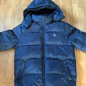 Svart vinterjacka från Calvin Klein - Snygg mörkblå pufferjacka från Calvin Klein Jeans med huva och dragkedja framtill. Jackan har diskret CK-logga på bröstet och ärmarna, samt två fickor med knappstängning. Perfekt för kalla dagar och riktigt stilren look.