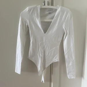 Vit långärmad korsett från Babaton - Säljer en vit långärmad korsett-topp från Babaton med djup v-ringning och spetsig nederkant. Toppens material är mjukt och stretchigt, perfekt för att skapa en snygg siluett. Stängs med knappar i grenen. Supersnygg att styla med jeans eller kjol! Shoulder pads