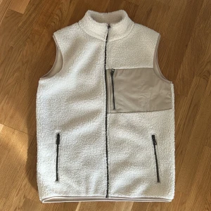 Pile väst  - Mysig vit fleeceväst med hög krage och hel dragkedja framtill. Västen har två sidofickor med dragkedja och en beige bröstficka med dragkedja. Materialet är fluffigt och ger en skön känsla, perfekt för lager-på-lager. Enkel och stilren design utan mönster.