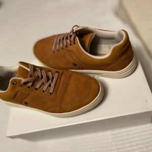 Bruna sneakers från Spanning Originals - Snygga bruna sneakers från Spanning Originals med beige sula och matchande snörning. Har använt de få gånger.Skorna har rund tå, diskreta detaljer och är tillverkade i mocka-liknande material. Perfekta för dig som gillar stilrena och bekväma skor med en urban vibe.