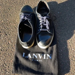 Svarta sneakers från Lanvin Paris - Snygga svarta sneakers från Lanvin Paris med klassisk design. Skorna har mörk blå snören, rund tå med lackad tåhätta och ovandel i mocka och skinn. Vit sula som ger en schysst kontrast. Perfekta för dig som gillar stilrena och exklusiva sneakers. Använda men fortfarande i bra skick, hör av dig vid minsta fundering! Kom gärna med bud. Det är en liten skada inne i skon vid hälen (se sista bilden)