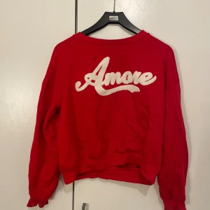 Röd sweatshirt med Amore-tryck - Cool röd sweatshirt med stort vitt Amore-tryck framtill. Tröjan har en avslappnad passform, rund halsringning och långa ärmar. Perfekt för dig som vill sticka ut med ett statement-plagg och gillar streetstyle.