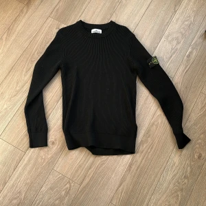 Svart stickad tröja från Stone Island i storlek L - Svart ribbstickad tröja från Stone Island med klassisk rund hals och ikonisk logotyp-patch på vänster ärm. Tröjan har lång ärm och är tillverkad i ett mjukt, stickat material som ger en clean och stilren look.