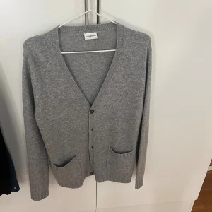 Grå kofta från John Henric - Klassisk grå kofta från John Henric i mjuk ullblandning. V-ringad med knappar framtill och två praktiska fickor. Perfekt för lager-på-lager och enkel att matcha med allt. Långärmad och ribbade muddar för extra komfort.