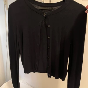 Svart kofta med knappar - En stilren svart kofta med rund halsringning och knäppning framtill. Koftan har långa ärmar och en klassisk, enkel design som passar till det mesta. Materialet känns mjukt och lätt