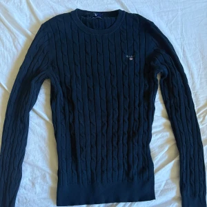 Mörkblå kabelstickad tröja från GANT - Kabelstickad mörkblå tröja från GANT i stretchig bomull. Klassisk rund halsringning och broderad GANT-logga på bröstet. Perfekt för dig som gillar preppy stil och vill ha något snyggt och bekvämt till vardags.