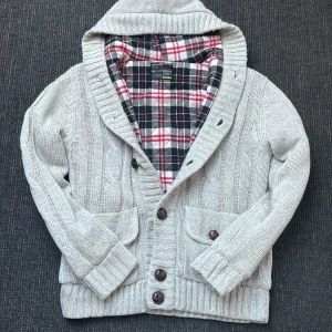 Stickad beige kofta  - Mysig stickad kofta från Brave Soul i beige med grova kabelstickade detaljer, stora bruna knappar och två fickor framtill. Koftan har huva och rutigt innerfoder i rött, vitt och svart. Perfekt för kyliga dagar och enkel att styla till jeans. Jätte bra skick. I storlek L hör av er vid funderingar!