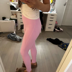 Ljusrosa höga leggings - Säljer ett par ljusrosa leggings med hög midja. De är tighta och stretchiga, perfekta för träning eller chill. Materialet känns mjukt och följsamt mot huden. Snygg passform som framhäver benen och midjan. Från Lululemon. Utan lapp då den störde mig. Kan leta efter orderbevis om de behövs.