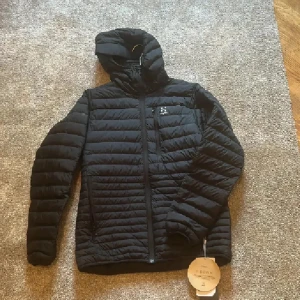 Svart dunjacka med huva från Haglöfs - Svart dunjacka från Haglöfs i modellen Micro Nordic Down Hood. Jackan har quiltad design, huva, dragkedja framtill och två sidofickor med dragkedja. Perfekt för kalla dagar och riktigt lätt att bära. Klassisk och stilren look.