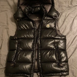 Svart dunväst från Moncler - Svart dunväst från Moncler med glansig finish, hög krage och avtagbar huva. Västen har två dragkedjeförsedda fickor och klassisk Moncler-logga på bröstet. Insidan har ett unikt serietidningsmönster och märkestaggar. Perfekt för lager-på-lager under kyliga dagar.