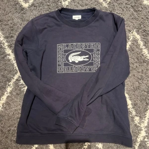 Lacoste tröja - Säljer en mörkblå långärmad tröja från Lacoste med tryckt logga och krokodil på bröstet samt broderad krokodil bak i nacken. Tröjan är i mjuk bomull och har en klassisk passform med rund hals.