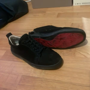 Christian Louboutin svarta sneakers - Svarta sneakers från Christian Louboutin med klassisk röd sula och signaturdetalj på hälen. Skorna har snörning, rund tå och är tillverkade i mocka. Box och dustbag medföljer. Perfekta för dig som vill sticka ut med exklusiva detaljer. Det finns ett hål därbk, därför de låga priset, skriv vid frågor, pris kan diskuteras vid snabb affär 