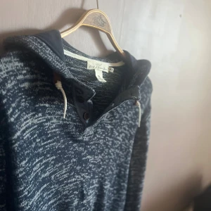 Mörkblå hoodie med snörning - Säljer en mörkblå och vitmelerad hoodie från Chums i storlek L. Tröjan har huva med snörning, knappar vid halsen och ribbade muddar. Materialet är stickat och känns mjukt och skönt. Perfekt för dig som gillar avslappnad stil.
