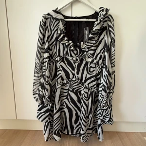 Zebramönstrad kortklänning från Boohoo - Snygg kortklänning från Boohoo med svartvitt zebramönster och volangdetaljer. Klänningen har långa ärmar med breda manschetter och en lätt v-ringning framtill. Luftigt material som ger en cool och trendig look.