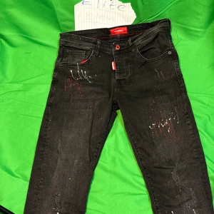 Svarta Dsquared2 ICON jeans med färgstänk - Snygga svarta jeans från Dsquared2 med stor 'ICON'-logga på bakfickan och coola färgstänk i vitt och rött framtill och baktill. Jeansen har klassisk femficksdesign, röda detaljer och smal passform. Perfekta för dig som vill sticka ut med en unik streetstyle.Liknande modell som originalet!