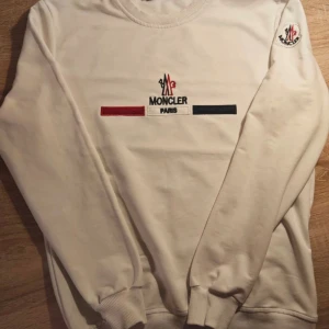 Vit Moncler  - Snygg vit sweatshirt från Moncler med logga och text på bröstet. Tröjan har rund hals, långa ärmar och ribbade muddar. Klassisk och stilren design med Moncler-märke även på ärmen.