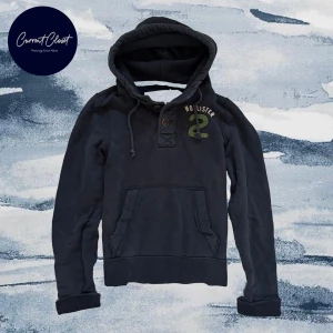 Mörkblå hoodie från Hollister - Mörkblå hoodie från Hollister med broderad grön siffra och logga på bröstet | Axelbredd: 41 cm längd: 62cm | Size M