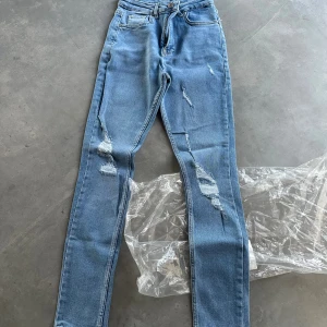 Ljusblå skinny jeans med slitningar XXS - Säljer ett par oanvända  ljusblå skinny jeans i storlek XXS med snygga slitningar på benen. Jeansen har klassisk femficksdesign, hög midja och är tillverkade i bomull med lite elastan för extra stretch. Perfekta för dig som gillar en trendig och avslappnad look.