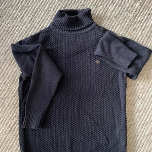 Mörkblå stickad polotröja Morris - Säljer en mörkblå stickad polotröja från Morris med snygg strukturstickning framtill och ribbade muddar. Klassisk polokrage och diskret Morris-logga på bröstet. Perfekt för dig som gillar stilrena och tidlösa plagg.